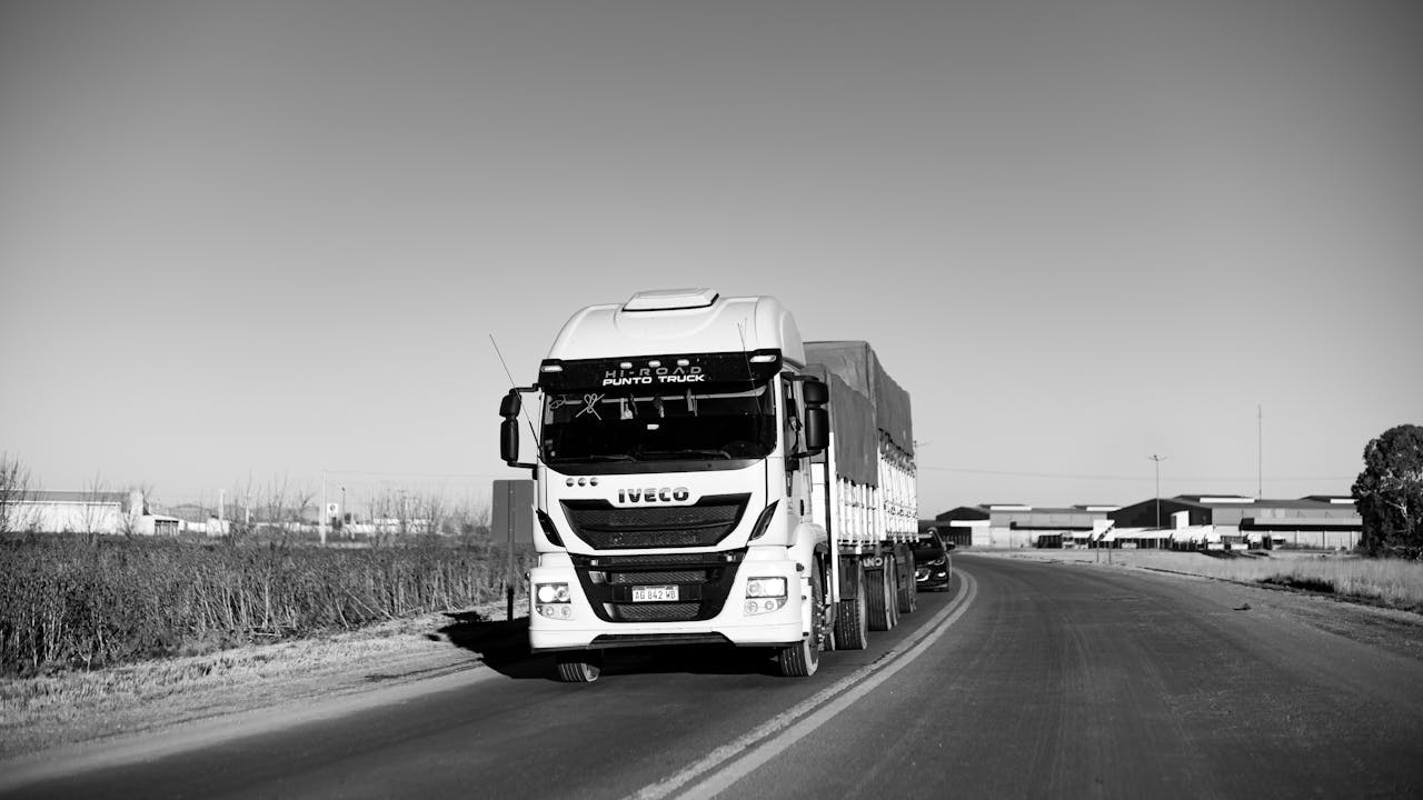 Monochrome image of a truck driving on a rural highway, emphasizing urban life.